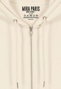 Felpa con zip in cotone crema, dotata di cerniera argentata, lacci e etichetta stampata. Presenta polsini e orlo a costine. Design semplice senza motivi.