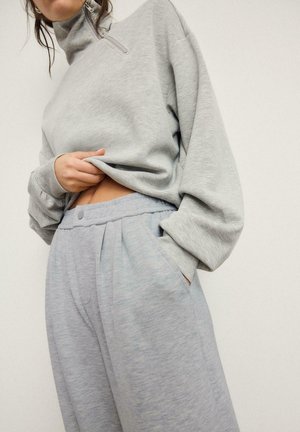 Frau trägt hellgraue, weit geschnittene Jogginghose und passenden Reißverschluss-Sweatshirt, eine Hand in der Tasche, die andere hebt das Sweatshirt leicht an.