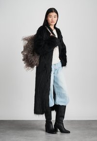 Frau in einem langen schwarzen Kunstfellmantel, hellblauen, weiten Jeans, schwarzen Stiefeln mit Absatz und mit einem durchsichtigen, gerafften braunen Rucksack.