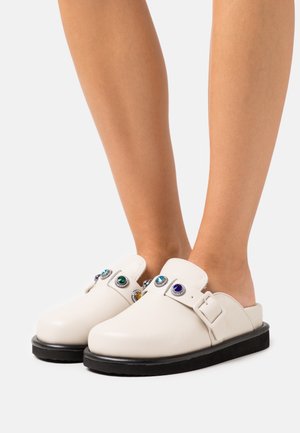 Sabots en cuir blanc avec un bout arrondi, ornés de plusieurs bijoux colorés et d'un détail de boucle. La semelle en caoutchouc noir apporte un contraste.