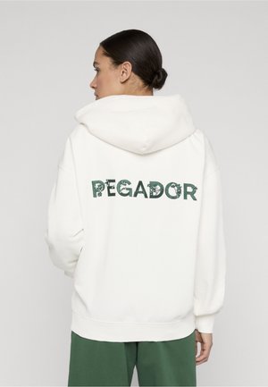 Kvinde med mørkt hår i en lav knold, iført en hvid hættetrøje med grøn blomstermønster og teksten "PEGADOR" på ryggen samt grønne bukser, set bagfra.