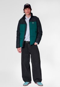 Kurtka puchowa z zielonym przodem i czarnymi rękawami, zestawiona z jasnoniebieską koszulą i szerokimi czarnymi dżinsami. Niebieskie buty i czarny kapelusz.