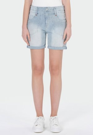 Vrouw draagt lichtblauwe en witte verticale gestreepte denimshorts met opgerolde boorden en witte sneakers, staand tegen een effen achtergrond.