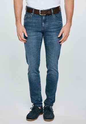 Mężczyzna w niebieskich dopasowanych dżinsach, brązowym skórzanym pasku i ciemnoniebieskich butach sznurowanych, stojący na jasnym, jednolitym tle.