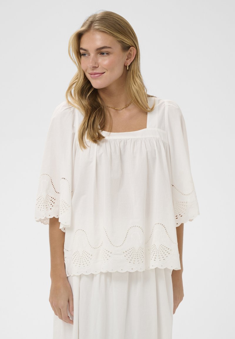 Femme blonde portant un chemisier blanc ample avec des motifs en œillets sur les manches et l'ourlet, souriante et regardant vers la gauche.