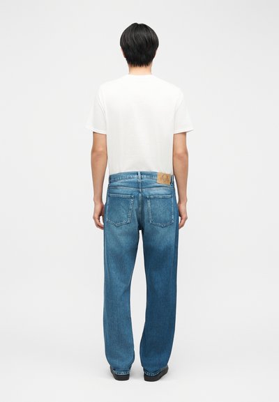 Άνδρας που στέκεται με την πλάτη γυρισμένη, φορώντας απλό λευκό μπλουζάκι, φαρδιά μπλε τζιν με πίσω τσέπες και μαύρα παπούτσια σε λευκό φόντο.