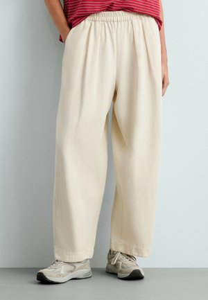 Personne portant un pantalon ample couleur crème et des baskets beiges, debout contre un mur uni de couleur claire, les mains dans les poches.