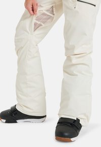 Cremefarbene Skihosen mit Reißverschlusstasche und Netzfutter, kombiniert mit schwarzen Snowboardstiefeln und weißen Sohlenakzenten.