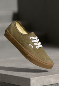 Olijfgroen suède sneaker met witte veters, bruine rubberen zool en Vans-logo. Eenvoudig ontwerp, lage silhouet, gladde textuur.