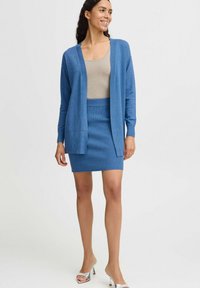 Lichtblauwe cardigan met een open voorkant, gecombineerd met een geribbelde lichtblauwe rok. Het materiaal lijkt zacht en lichtgewicht. Zilveren hoge hakken.