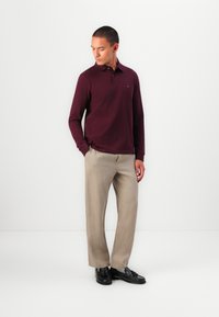 Burgundy long-sleeve polo shirt with a classic collar, paired with light beige trousers and black loafers; smooth fabric and relaxed fit.
