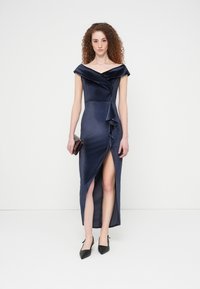 Φόρεμα σε ναυτικό μπλε χρώμα με ανοιχτούς ώμους από υφασμάτινο υλικό με υφή, με λεπτομέρεια βολάν στο μπροστινό μέρος και πλαϊνή σχισμή, συνδυασμένο με μαύρες μυτερές γόβες.