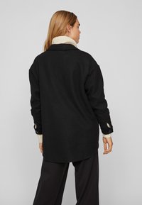 Črni oversize plašč s ovratnikom, ki ima teksturirano tkanino, gumbaste manšete in pod njim kremo pulover s turtleneck ovratnikom.