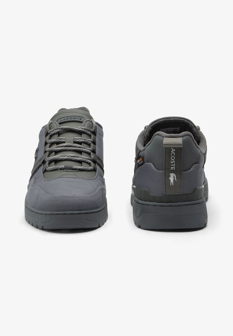 Paire de baskets grises Lacoste, une montrant l’avant avec des lacets, l’autre montrant l’arrière avec le logo et la languette sur fond blanc.