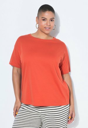 Woman with short hair wearing gold hoop earrings, an orange short-sleeve top, and black-and-white striped pants, standing against a white background.