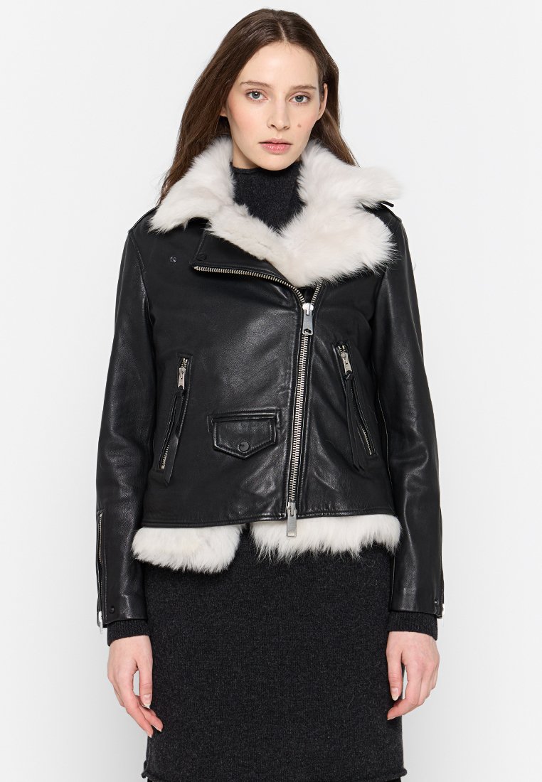 Eine Frau trägt eine schwarze Lederjacke mit weißem Kunstfellkragen und -saum und steht vor einem schlichten weißen Hintergrund.