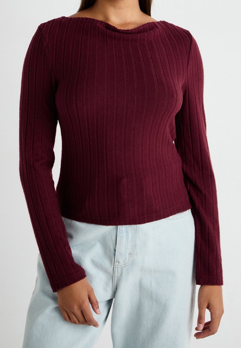 Personne portant un pull ajusté en maille côtelée bordeaux à manches longues et un jean taille haute bleu clair, debout devant un fond uni.