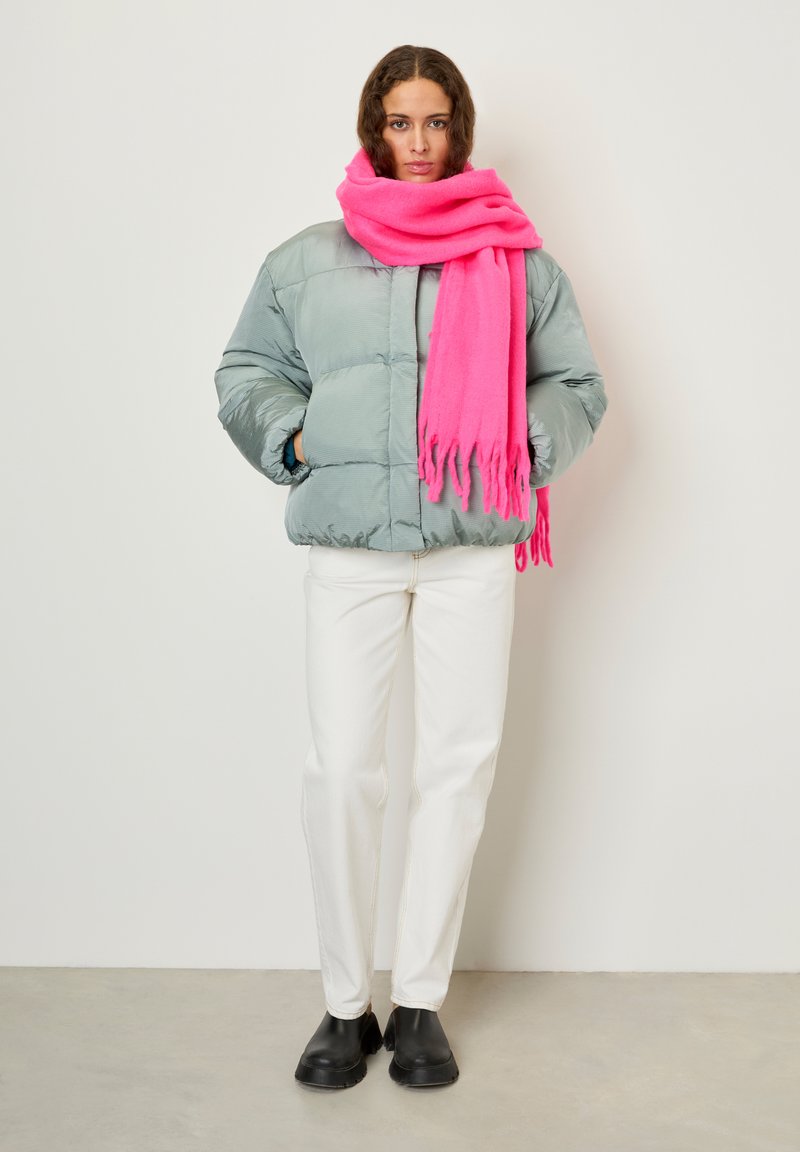 Woman wearing a light blue puffer jacket, bright pink scarf, white pants, and black boots standing against a plain background.
