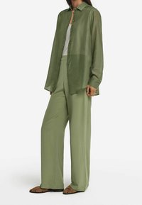Chemise boutonnée verte légère et semi-transparente portée sur un débardeur blanc, associée à un pantalon vert ample et des sandales marron.