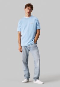 T-shirt azzurro chiaro a maniche corte con collo rotondo e piccolo logo sul petto, abbinato a jeans blu sbiaditi e sneaker bianche.
