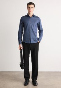 Cămașă albastră în dungi, cu mânecă lungă și guler ascuțit, purtată cu pantaloni negri și pantofi negri. Ținând o geantă de umăr neagră.