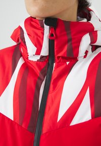 Veste rouge et blanche avec des motifs abstraits, dotée d'un col haut, d'une fermeture éclair sur le devant et d'une texture brillante. Accentuée par un tirette de fermeture contrastante.