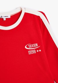 Rode t-shirt met een witte ronde hals en accentstrookjes op de mouwen. Bevat de tekst "SEIZOEN 1998" in het wit, met daaronder twee sterren.