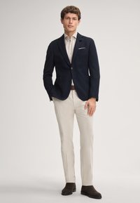 Blazer blu navy a righe con due bottoni, camicia bianca sotto, pantaloni beige e stivali alla caviglia marroni; design semplice con un fazzoletto da taschino.
