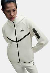 Sudadera con cremallera de Nike en blanco, con detalles en negro y un patrón distintivo. Tiene un alto cuello, cremallera completa y un bolsillo en la manga.