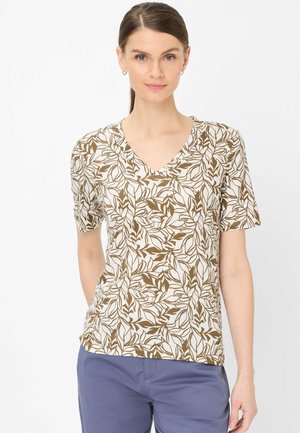 Frau mit glattem braunem Haar trägt weißes V-Ausschnitt-T-Shirt mit braunem Blattmuster und blaue Hose, steht vor weißem Hintergrund.