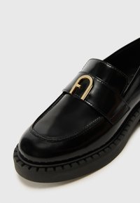 Black leather loafer with a rounded toe, featuring a gold-tone metal buckle accent. Thick black rubber sole with textured edges.