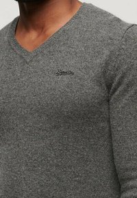 Pull en V gris en tissu tricoté. Avec un petit logo brodé sur la poitrine et une surface texturée. Silhouette ajustée.