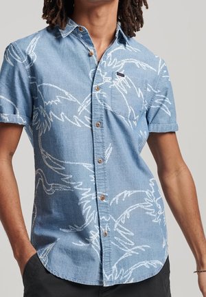 Homme portant une chemise bleu clair à manches courtes avec des motifs abstraits d'oiseaux blancs, debout les mains dans les poches.