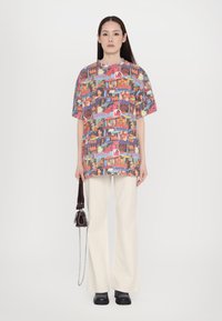 T-shirt z krótkim rękawem w oversize'owym fasonie, zdobiony kolorową grafiką w formie kolażu, w połączeniu z jasnobeżowymi spodniami w szerokim kroju i małą ciemną torebką.