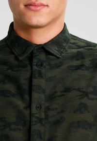 Ciemnozielona koszula zapinana na guziki z subtelnym wzorem kamuflażu, z klasycznym kołnierzem i czarnymi guzikami z przodu. Teksturowany materiał.