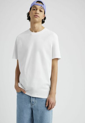 Jeune homme portant un t-shirt blanc, un jean bleu clair et une casquette de baseball lavande portée à l'envers, debout devant un fond uni.