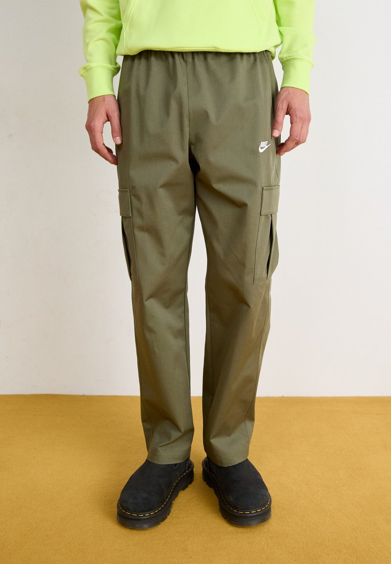 Ελιάς πράσινα cargo παντελόνια με ελαστική μέση, πλαϊνές τσέπες και λευκό λογότυπο. Φορεμένα με μαύρες σουέτ γόβες σε κίτρινο φόντο.