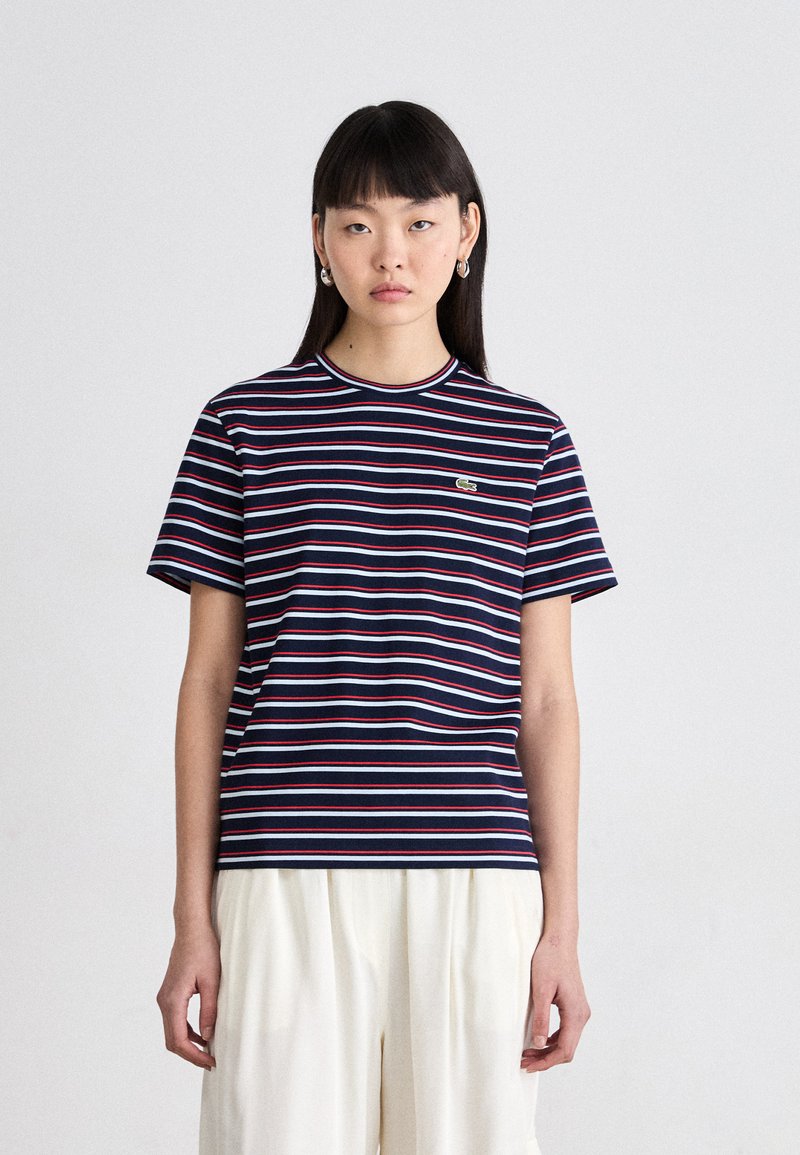 T-shirt rayé bleu marine avec des accents rouges et bleu clair, col rond, manches courtes, comportant un petit logo vert sur la poitrine.