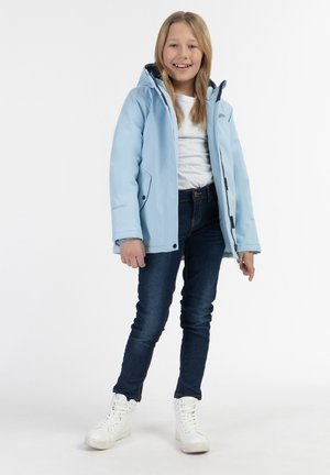 Lyseblå vandtæt jakke med hætte, lynlås og lommer, kombineret med mørke jeans og hvide sneakers. Pige, der står og smiler.