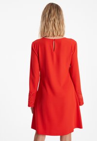 Rode jurk met lange mouwen. Bevat kanten accenten op de mouwen en een kleine opening aan de achterkant. Glad materiaal en A-lijn ontwerp.