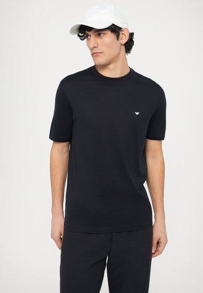 Čierne tričko s krátkym rukávom z bavlneného materiálu s okrúhlym výstrihom. Na ľavej strane na hrudi je malá biela logo. Nosí sa s tmavými textúrovanými nohavicami.