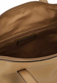Innenseite einer tan-farbenen Lederhandtasche mit einer weichen Textur, ausgestattet mit einem Reißverschlussfach und einem Etikett an der Seite. Glatte Stoffauskleidung im gesamten Innenraum.