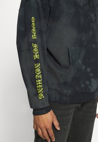 Tmavý mikina s tie-dye vzorem, má klokaní kapsu a žebrované manžety. Na rukávu je žlutý text. Vyrobena z měkké, strukturované tkaniny.