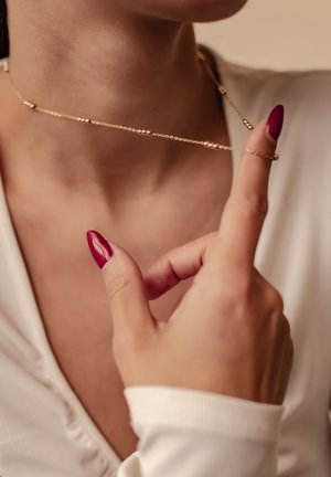 Gold chain necklace with small spherical beads, draped around the neck. A hand with red nail polish gently holds the necklace.