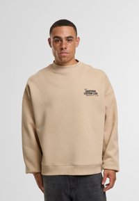 Felpa beige oversize con collo alto, spalle scese e un piccolo logo nero sul petto sinistro. Materiale in cotone morbido.