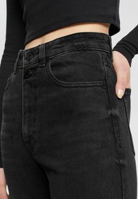 Jeans de mezclilla negra de tiro alto, con dos cierres de botón, bolsillos delanteros y una textura sutil. Vista en primer plano que muestra el ajuste y la tela.