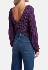 Maglione viola a maglia con texture ondulata, design con schiena bassa e maniche lunghe, abbinato a jeans skinny blu.