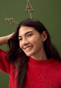 Gouden hoofdband met twee decoraties van draadbomen, versierd met groene en rode kralen, gedragen bij een rode pluche trui tegen een groene achtergrond.