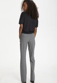 Camicia nera a maniche corte abbinata a pantaloni grigi su misura. I pantaloni hanno una texture liscia e tasche laterali. Il modello è di spalle rispetto alla fotocamera.