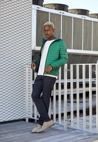 Grüne Daunenjacke mit horizontal gequilteter Textur, weißes T-Shirt, karierte marineblaue Hose und beige Schuhe, vor einem metallischen Hintergrund.